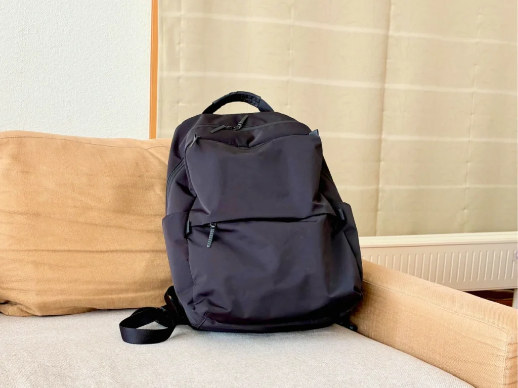 ソファ上に置いたファンクショナルバックパック正面。マットな生地が前面に広がり、中央に立体ポケット、上部に横ファスナー付き。